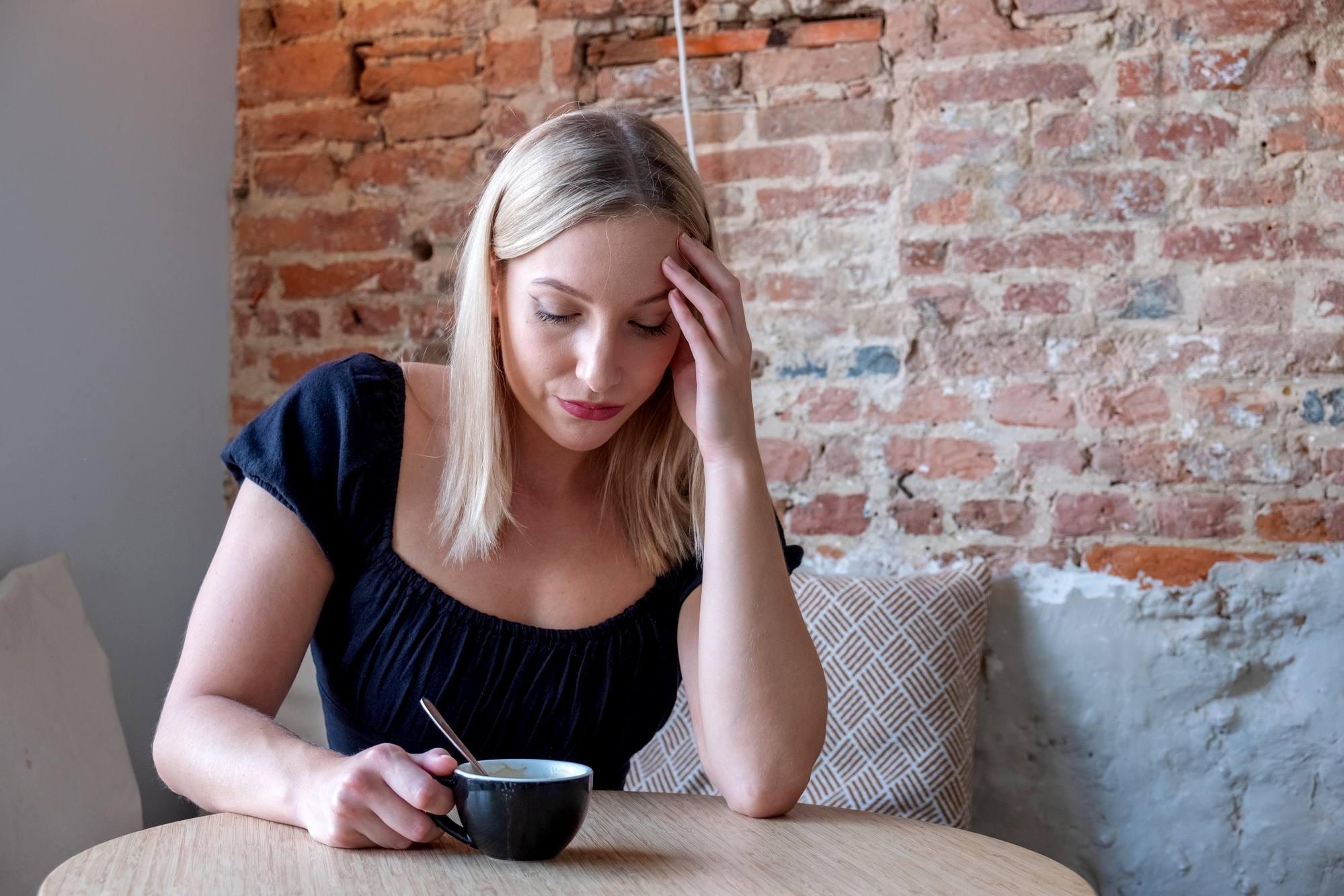 A woman with blonde hair sits at a wooden table in a cafe, holding a black coffee cup with a spoon inside. She rests her head on her hand, looking thoughtful with a pensive expression. The background features an exposed brick wall and a beige cushion.