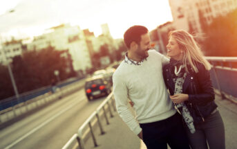 A couple walks on a bridge at sunset, smiling and holding each other closely. The man wears a white sweater and dark pants, while the woman wears a black jacket and scarf. Buildings and a few cars are visible in the background.