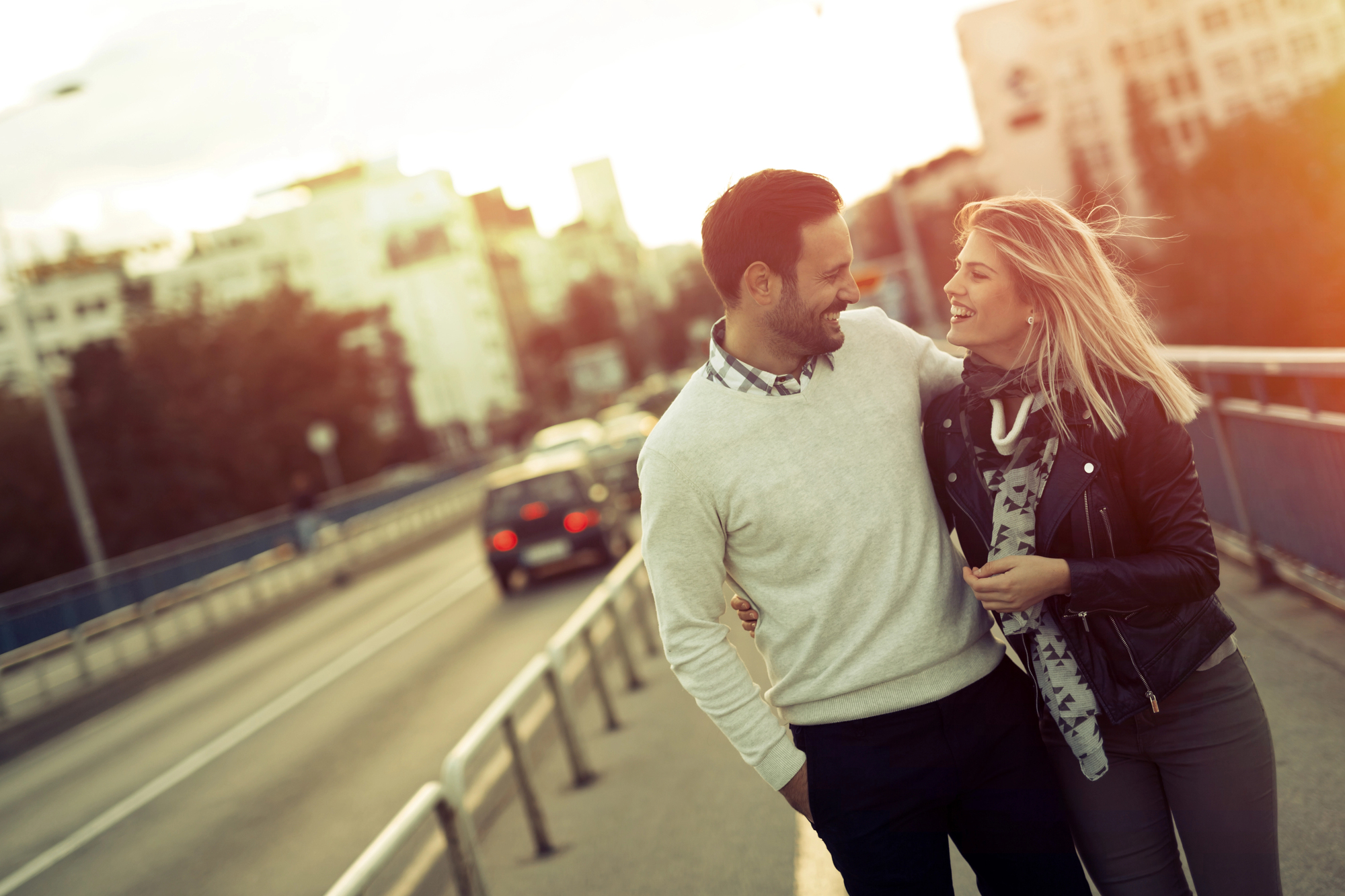 A couple walks on a bridge at sunset, smiling and holding each other closely. The man wears a white sweater and dark pants, while the woman wears a black jacket and scarf. Buildings and a few cars are visible in the background.