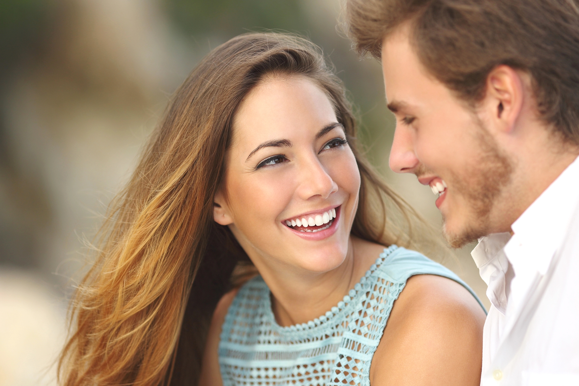 A woman with long brown hair and a man with short brown hair share a joyful moment, smiling at each other. The woman is wearing a sleeveless blue top, and the man is wearing a white shirt. The background is blurred, suggesting an outdoor setting.