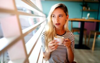 A young woman with long blonde hair sips from a white coffee cup while seated indoors near a large window with horizontal blinds. She is wearing a black and white striped shirt and looking pensively outside. The background shows a cafe interior with tables and chairs.