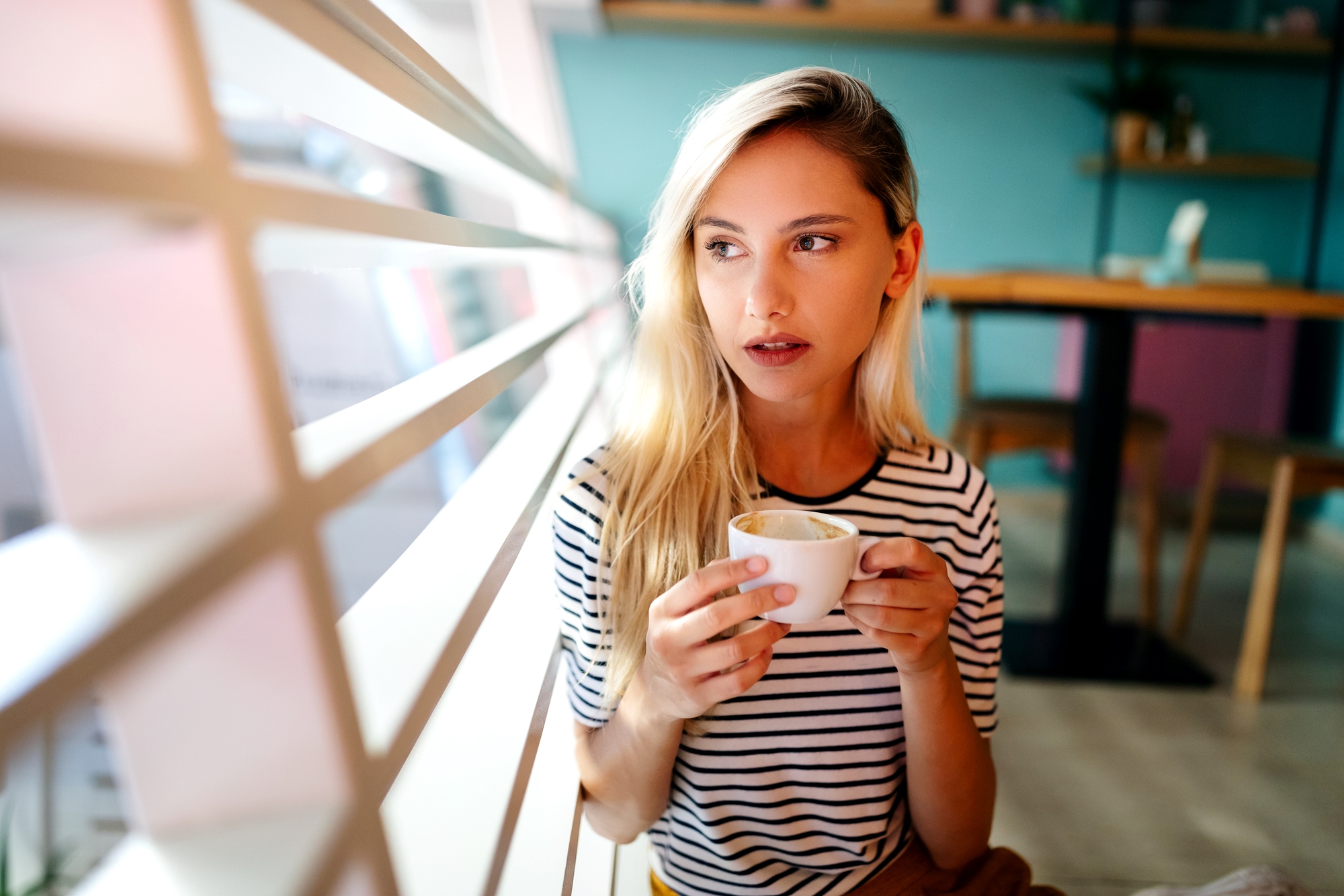 A young woman with long blonde hair sips from a white coffee cup while seated indoors near a large window with horizontal blinds. She is wearing a black and white striped shirt and looking pensively outside. The background shows a cafe interior with tables and chairs.