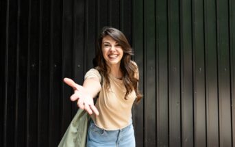A woman with long brown hair, wearing a beige t-shirt and jeans, smiles and reaches one hand towards the camera. She is standing in front of a black corrugated metal wall and has a light jacket draped over her shoulder.