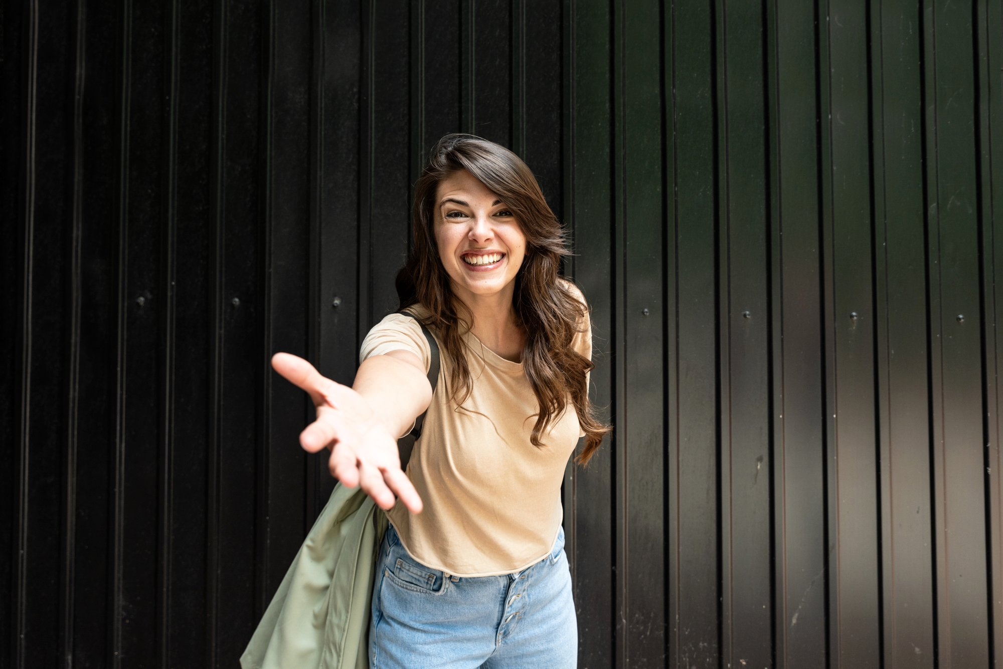 A woman with long brown hair, wearing a beige t-shirt and jeans, smiles and reaches one hand towards the camera. She is standing in front of a black corrugated metal wall and has a light jacket draped over her shoulder.