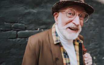 An elderly man with a white beard and glasses is smiling at the camera. He is wearing a plaid-patterned jacket, a white turtleneck, and a hat. The background is a brick wall.