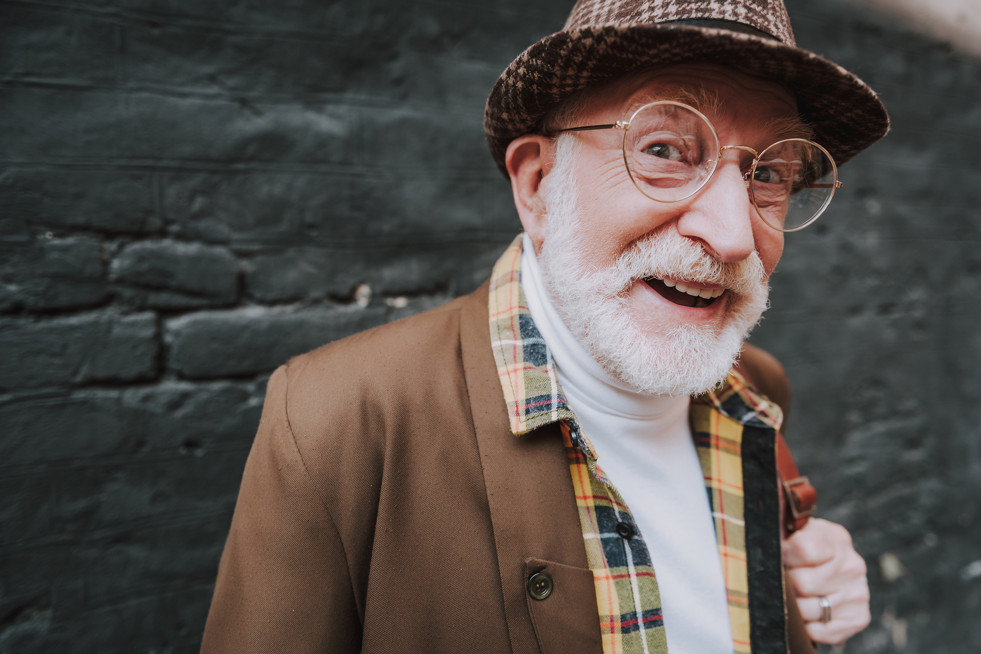 An elderly man with a white beard and glasses is smiling at the camera. He is wearing a plaid-patterned jacket, a white turtleneck, and a hat. The background is a brick wall.