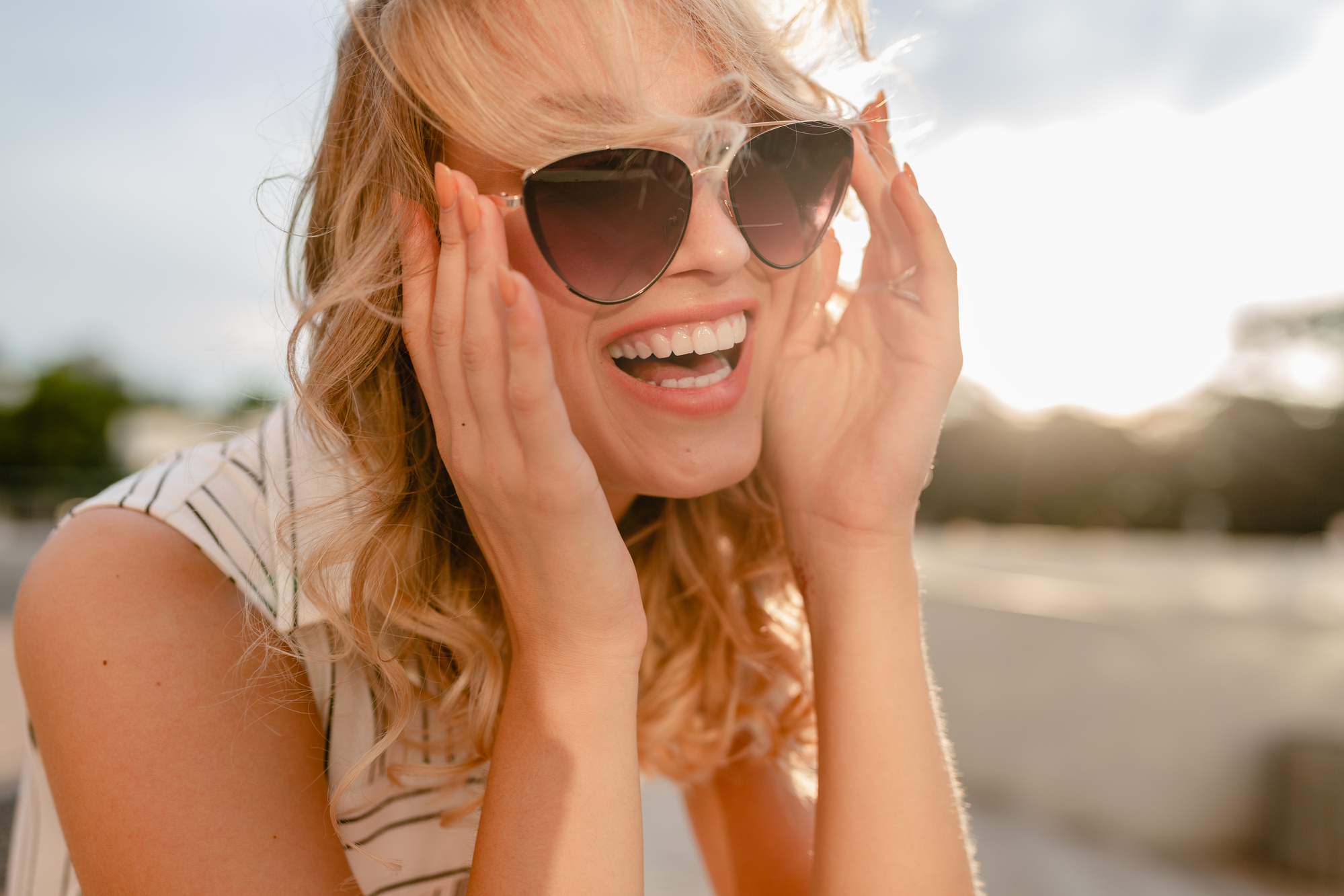 A smiling person with blonde, curly hair wearing sunglasses and a striped shirt, holds the sides of their sunglasses outdoors on a sunny day. The background shows a bright sky and some blurred greenery.