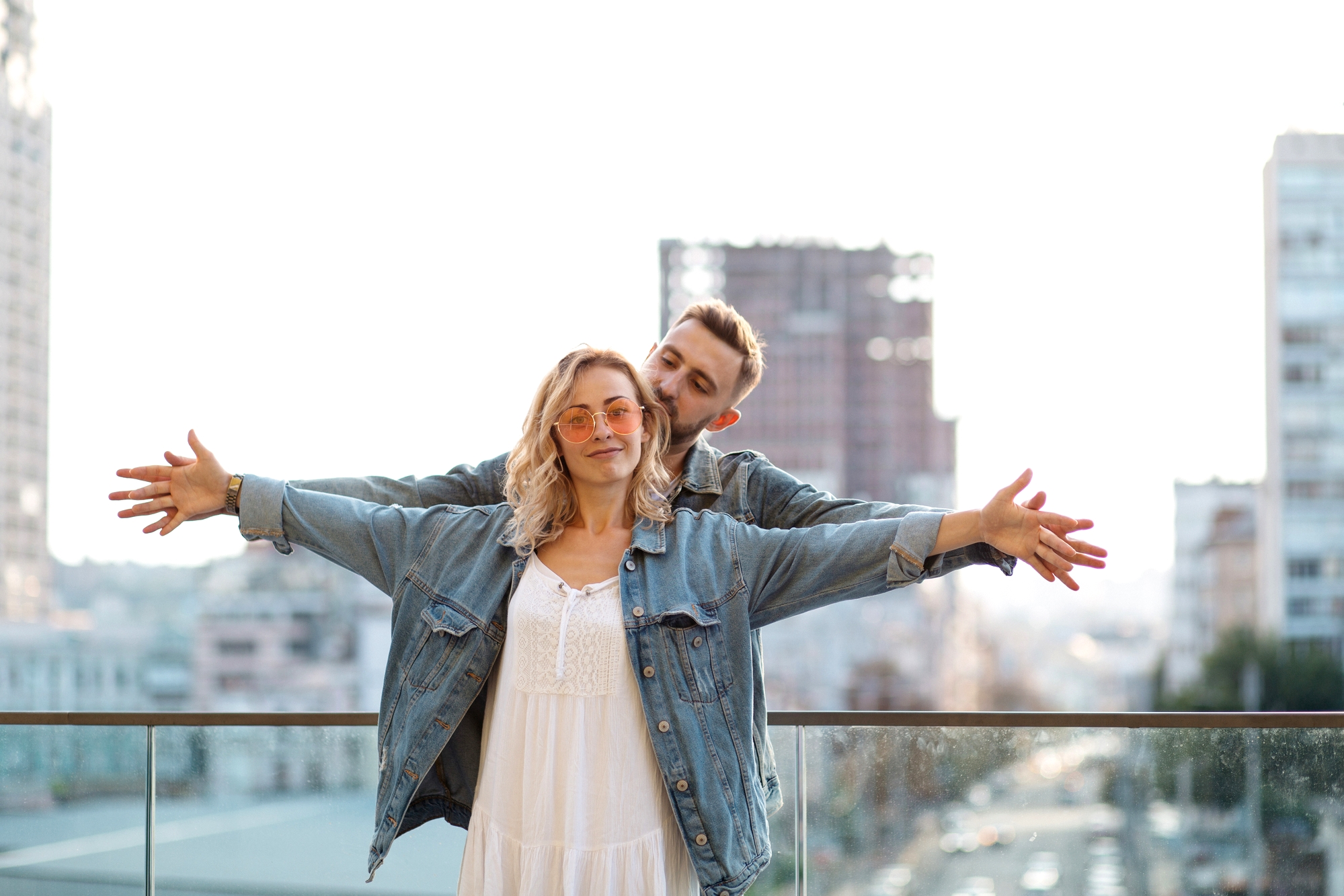 A couple stands on a rooftop with a cityscape in the background. The woman, wearing sunglasses and a white dress with a denim jacket, extends her arms wide, while the man, also in a denim jacket, stands behind her mimicking the same gesture and smiling.