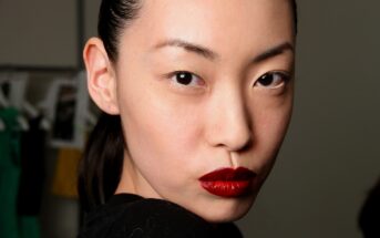 A woman with smooth skin, dark hair slicked back, and bold red lipstick looks directly at the camera with a serious expression. She is wearing a black top, and the background is slightly blurred, showing indistinct objects and some hanging clothing.