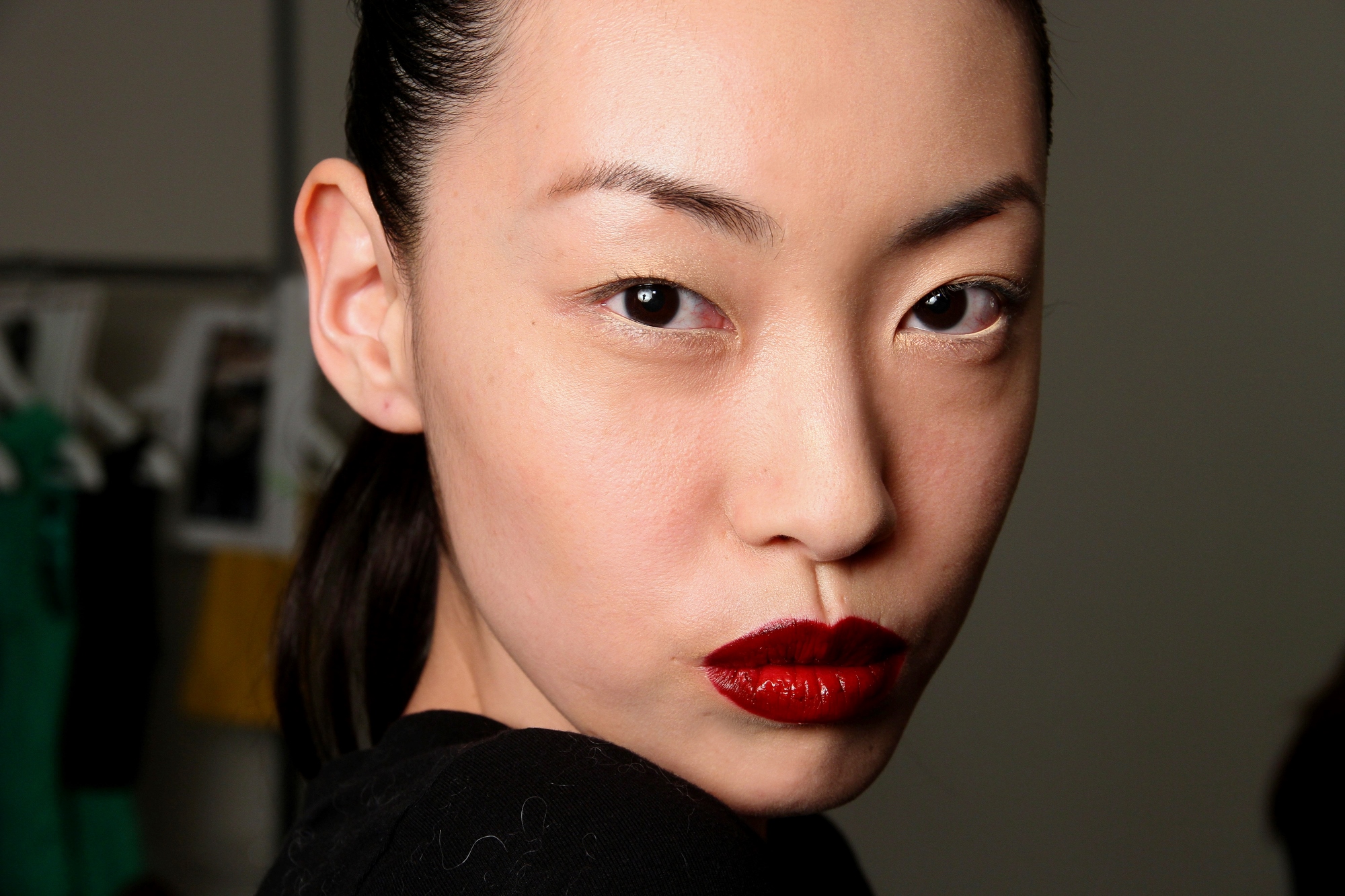 A woman with smooth skin, dark hair slicked back, and bold red lipstick looks directly at the camera with a serious expression. She is wearing a black top, and the background is slightly blurred, showing indistinct objects and some hanging clothing.