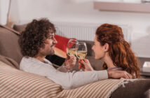A couple sits on a cozy couch, facing each other and smiling, as they clink glasses of white wine. The setting appears warm and comfortable, with a blanket draped over the sofa and soft lighting creating a relaxed atmosphere.