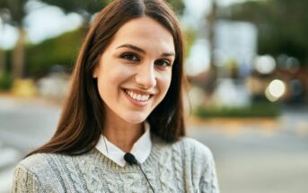 A young woman with long brown hair smiles at the camera. She is wearing a gray knitted sweater over a white collared shirt. The background is an outdoor urban setting with blurred trees and a building. The lighting suggests it is daytime.