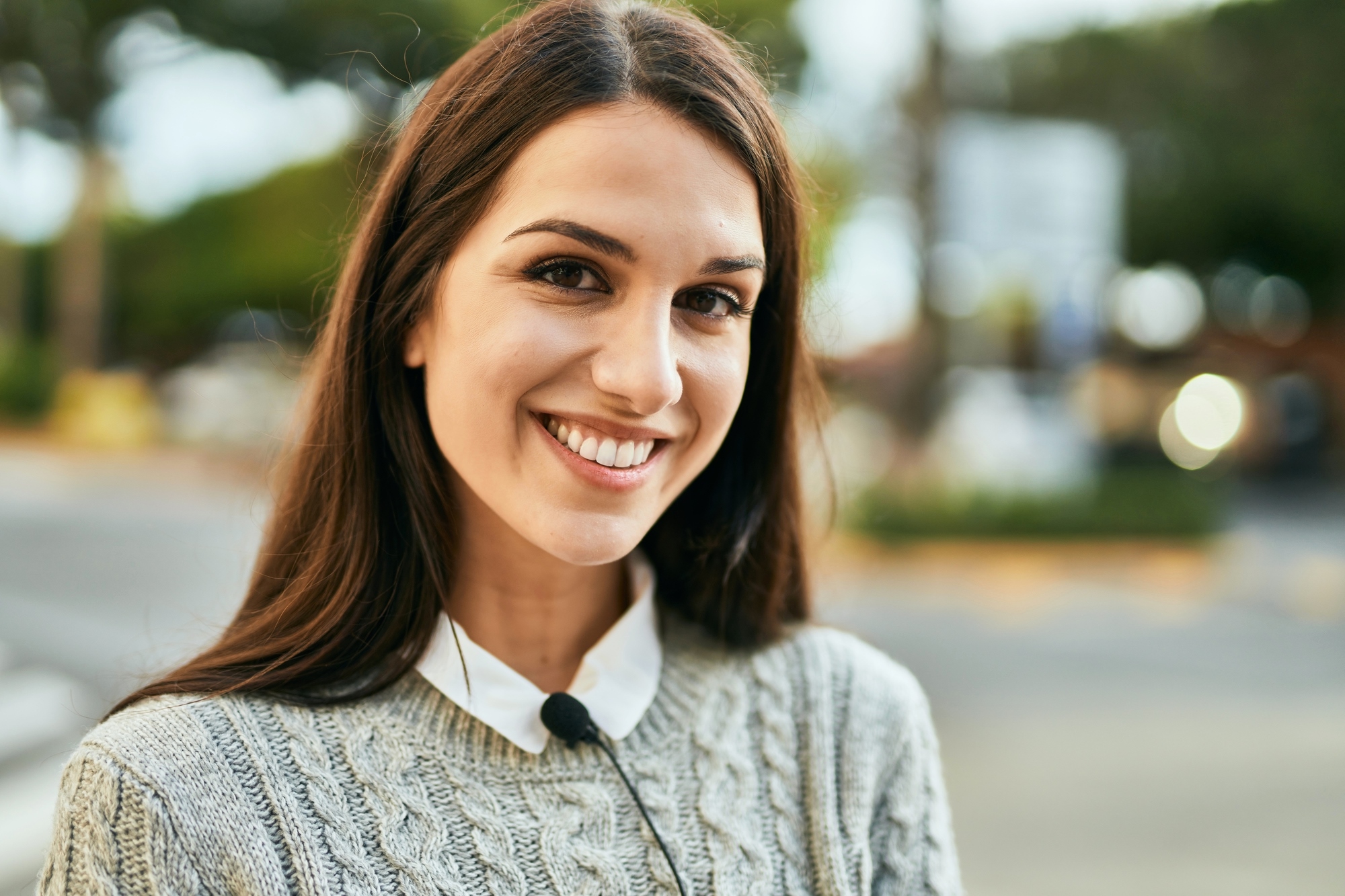 A young woman with long brown hair smiles at the camera. She is wearing a gray knitted sweater over a white collared shirt. The background is an outdoor urban setting with blurred trees and a building. The lighting suggests it is daytime.