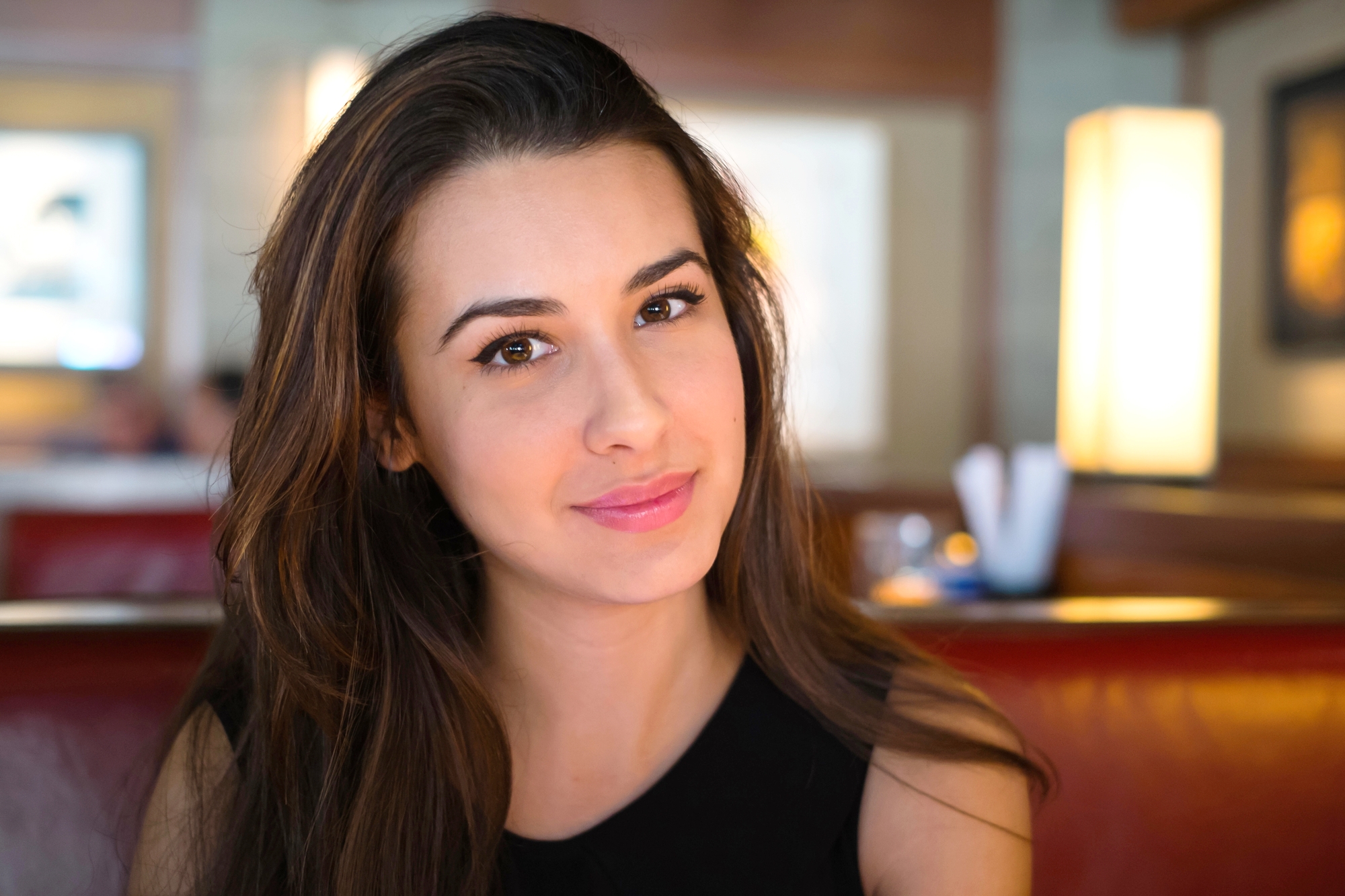 A woman with long, brown hair and a slight smile is sitting indoors. She is wearing a sleeveless black top. The background is softly blurred, showing a warm, well-lit interior with hanging lights and some blurred figures in the distance.