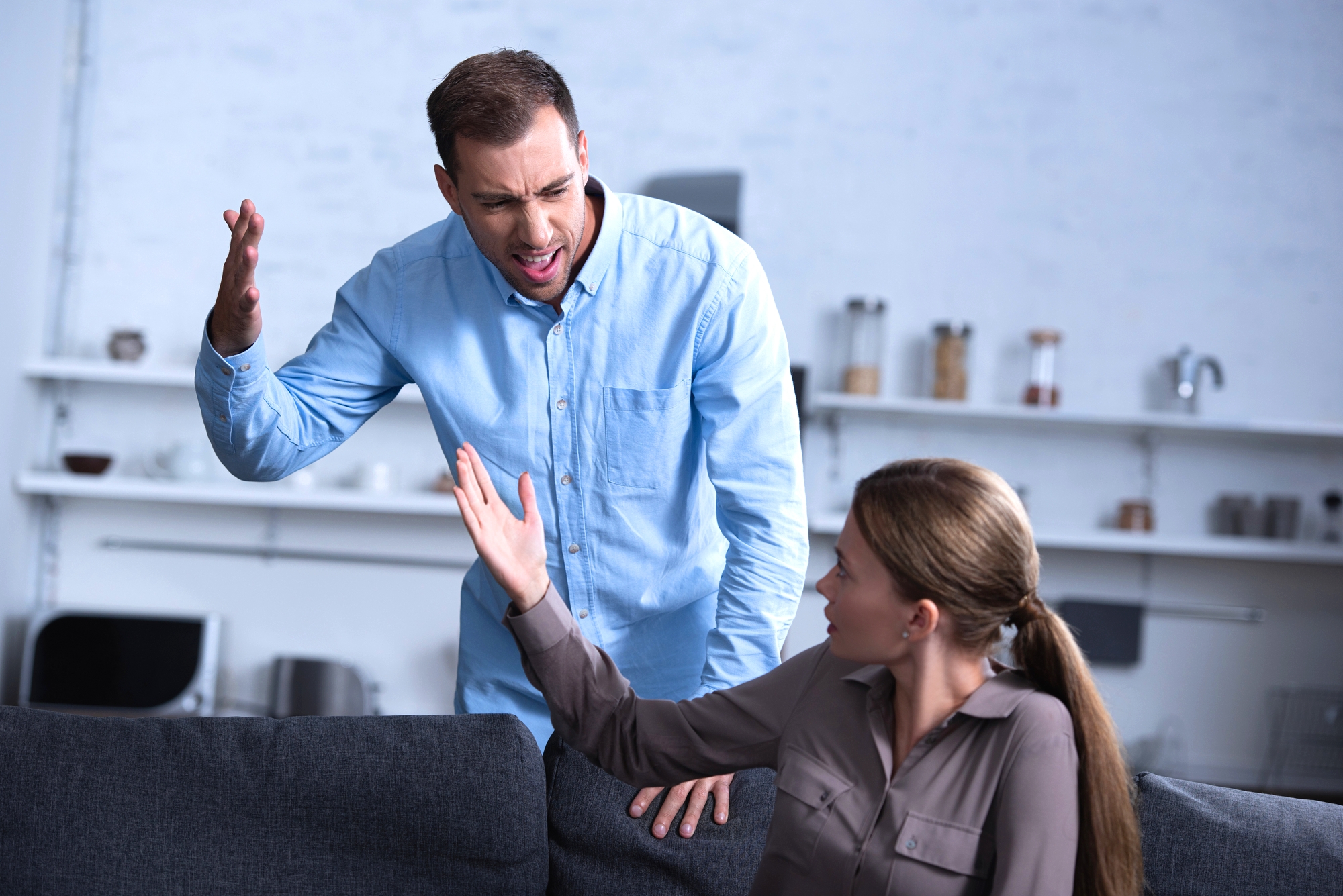 A man and a woman are having an intense argument in a living room. The man, standing and dressed in a blue shirt, is raising his hand and appears frustrated. The woman, sitting on a couch and wearing a gray shirt, is gesturing back at him, looking upset.