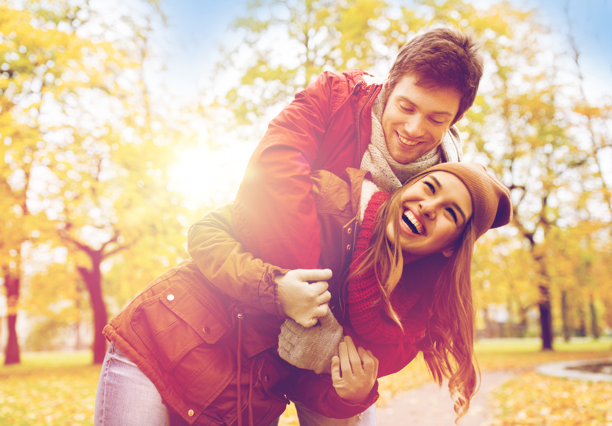 A young couple dressed in autumn clothing is playfully embracing in a park surrounded by colorful fall foliage. The man, in a red jacket, is leaning over the smiling woman, who is wearing a hat and scarf. They appear happy and joyful in the sunny, vibrant setting.