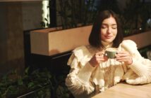 A woman with shoulder-length dark hair, wearing a cream-colored, ruffled sweater, sits at a wooden table in a cozy, plant-filled café. She holds a green mug with both hands, her eyes closed, and appears to be savoring the warmth and aroma of her drink.