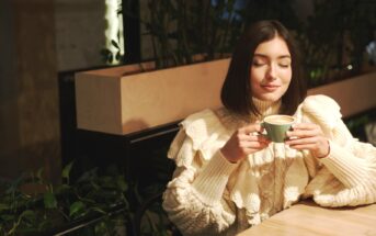 A woman with shoulder-length dark hair, wearing a cream-colored, ruffled sweater, sits at a wooden table in a cozy, plant-filled café. She holds a green mug with both hands, her eyes closed, and appears to be savoring the warmth and aroma of her drink.