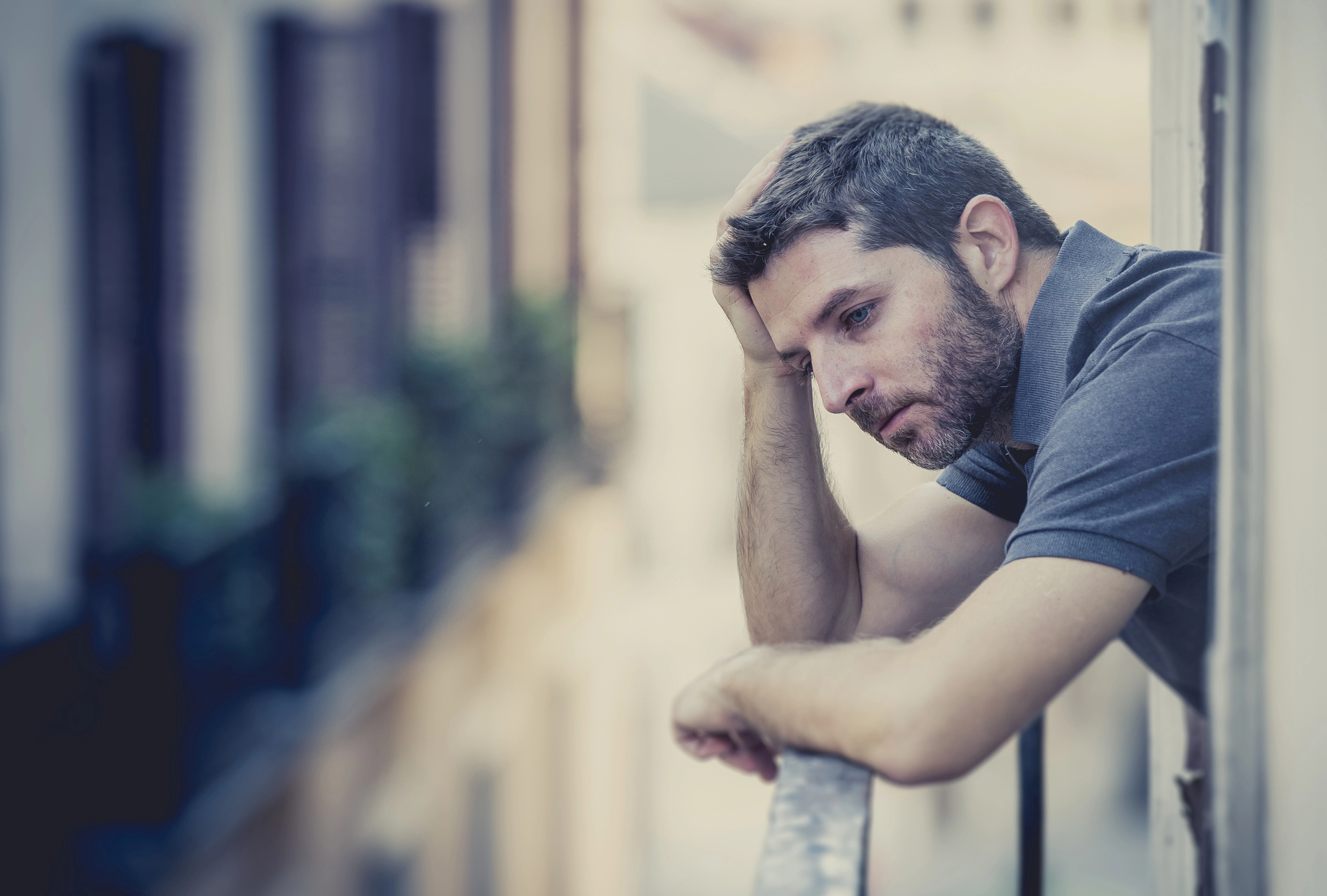 A man with short hair and a beard, wearing a gray polo shirt, rests his head on his hand while leaning on a balcony railing. He looks thoughtfully into the distance, with a blurred background of buildings and greenery.