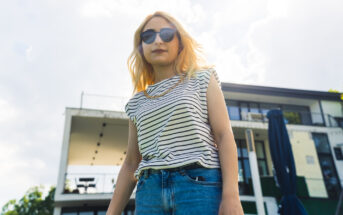 A woman with long blonde hair wearing sunglasses and a striped shirt stands confidently outdoors. She has one hand in the pocket of her blue jeans. In the background, there is a modern building with large windows and a patio area. The sky is partly cloudy.