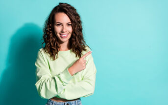 A woman with wavy brown hair smiles while pointing to the right. She is wearing a light green sweater and jeans. The background is a solid light blue color.