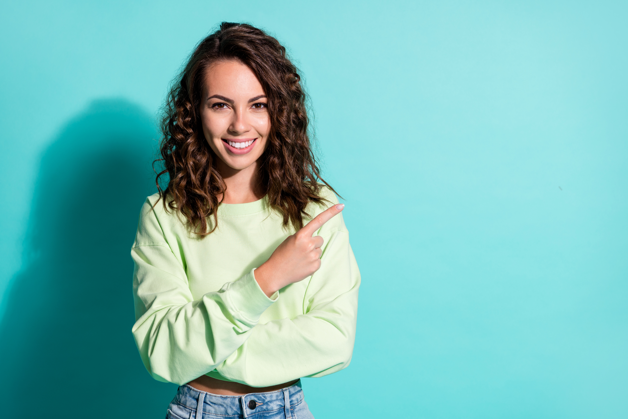 A woman with wavy brown hair smiles while pointing to the right. She is wearing a light green sweater and jeans. The background is a solid light blue color.