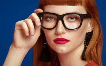 A person with long red hair and blunt bangs wears large black glasses, bold red lipstick, and black tassel earrings. They have a serious expression and are posing against a dark blue background, with their hand resting near their cheek, touching the glasses.