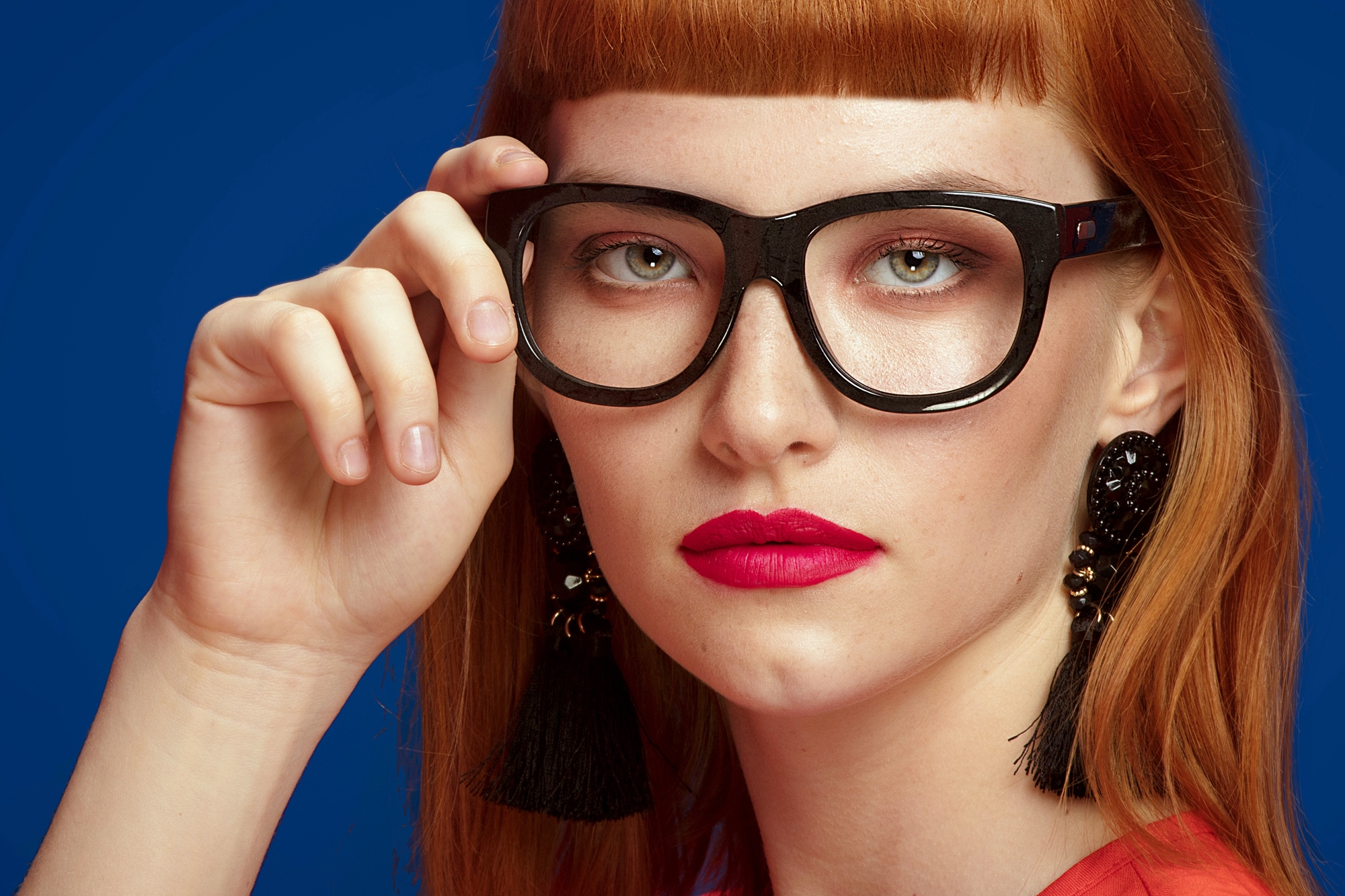 A person with long red hair and blunt bangs wears large black glasses, bold red lipstick, and black tassel earrings. They have a serious expression and are posing against a dark blue background, with their hand resting near their cheek, touching the glasses.