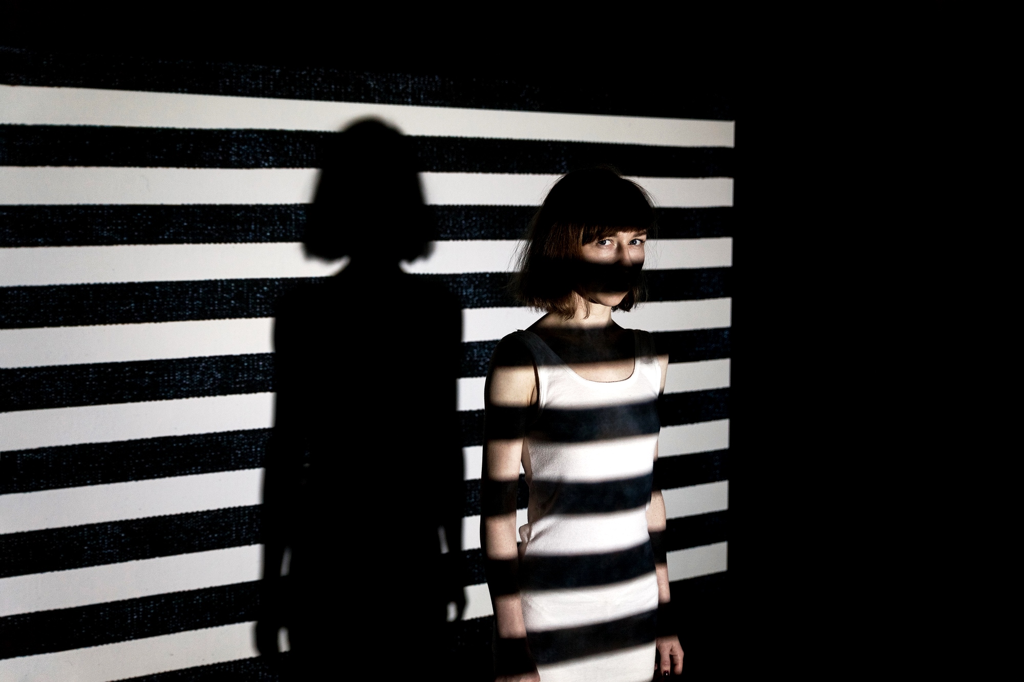 A woman stands against a background with horizontal black and white stripes. The lighting creates a shadow of the stripes across her face and body, blending her partially into the backdrop. Her full shadow is projected onto the striped wall.