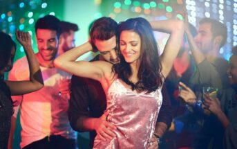 A group of people are dancing at a lively party with colorful lights in the background. At the center, a smiling woman in a pink velvet top dances closely with a man behind her. Others around them are also enjoying the music and atmosphere.