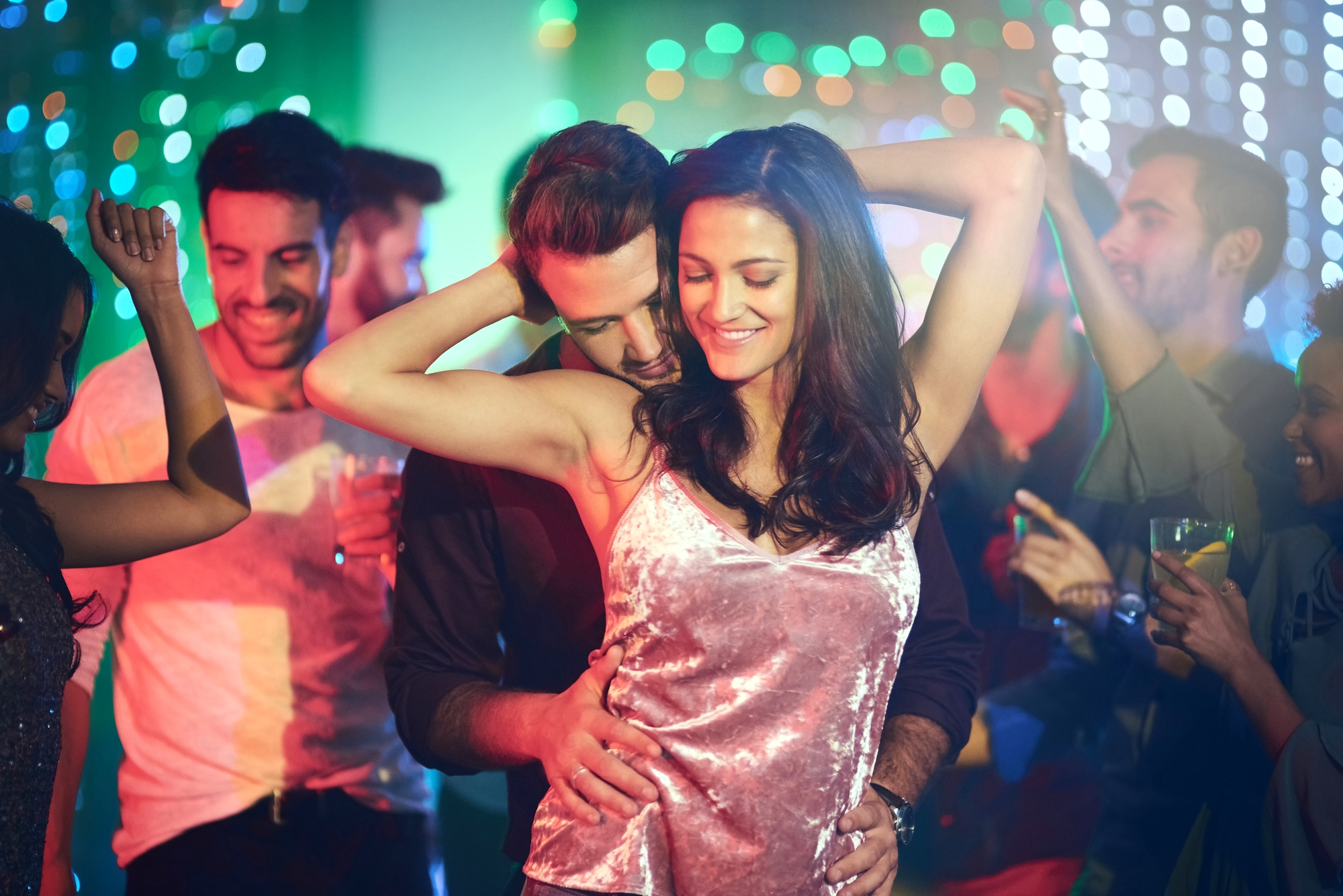 A group of people are dancing at a lively party with colorful lights in the background. At the center, a smiling woman in a pink velvet top dances closely with a man behind her. Others around them are also enjoying the music and atmosphere.