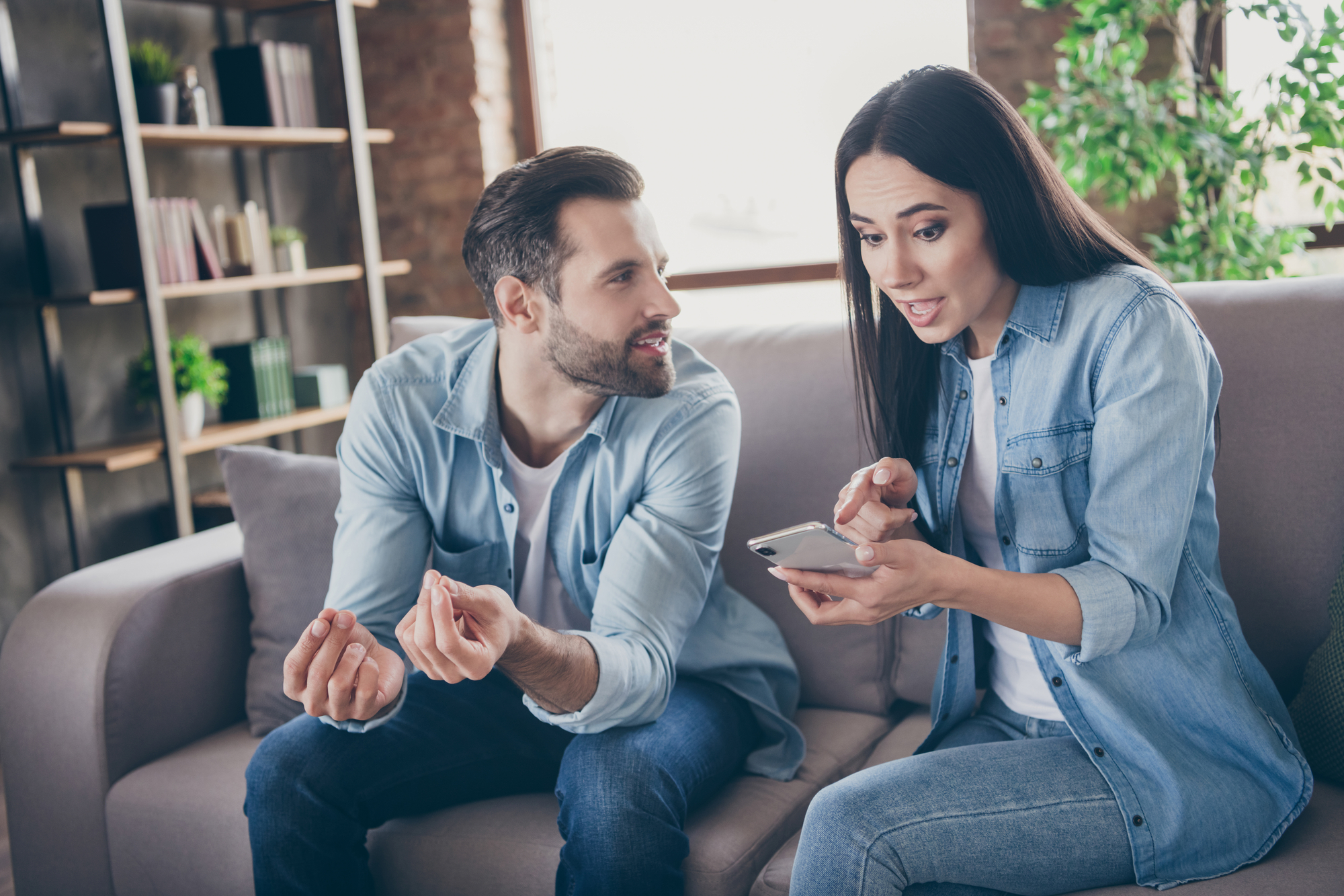 A man and a woman sit on a couch, both dressed in casual denim shirts. The woman is looking surprised while holding a smartphone, showing something to the man, who leans in with a curious expression. A bookshelf with decor is in the background.