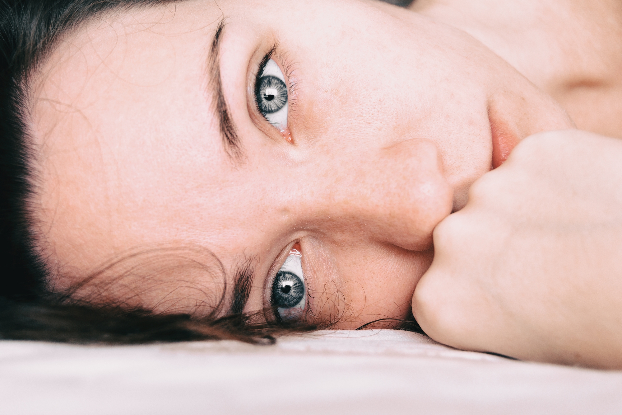 Close-up of a person's face lying down with their cheek resting on a surface. The individual's blue eyes are gazing intently towards the camera, and their hand is partially covering the lower half of their face.