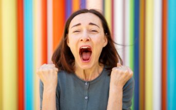 A woman with shoulder-length brown hair passionately shouts with both fists clenched in front of a vibrant, colorful striped background. She appears to be celebrating or expressing excitement. She is wearing a green top.