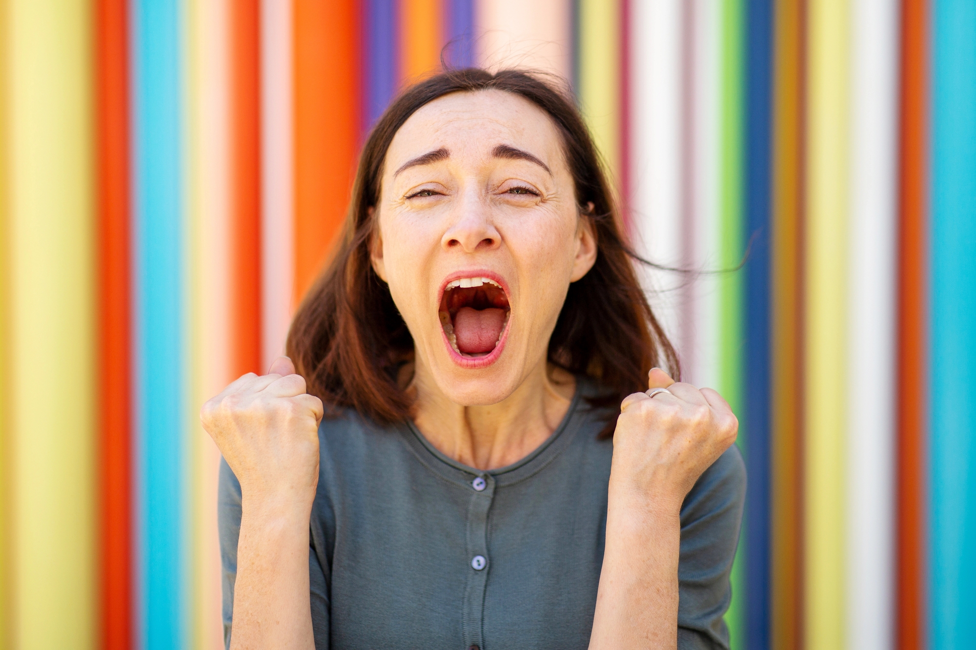 A woman with shoulder-length brown hair passionately shouts with both fists clenched in front of a vibrant, colorful striped background. She appears to be celebrating or expressing excitement. She is wearing a green top.