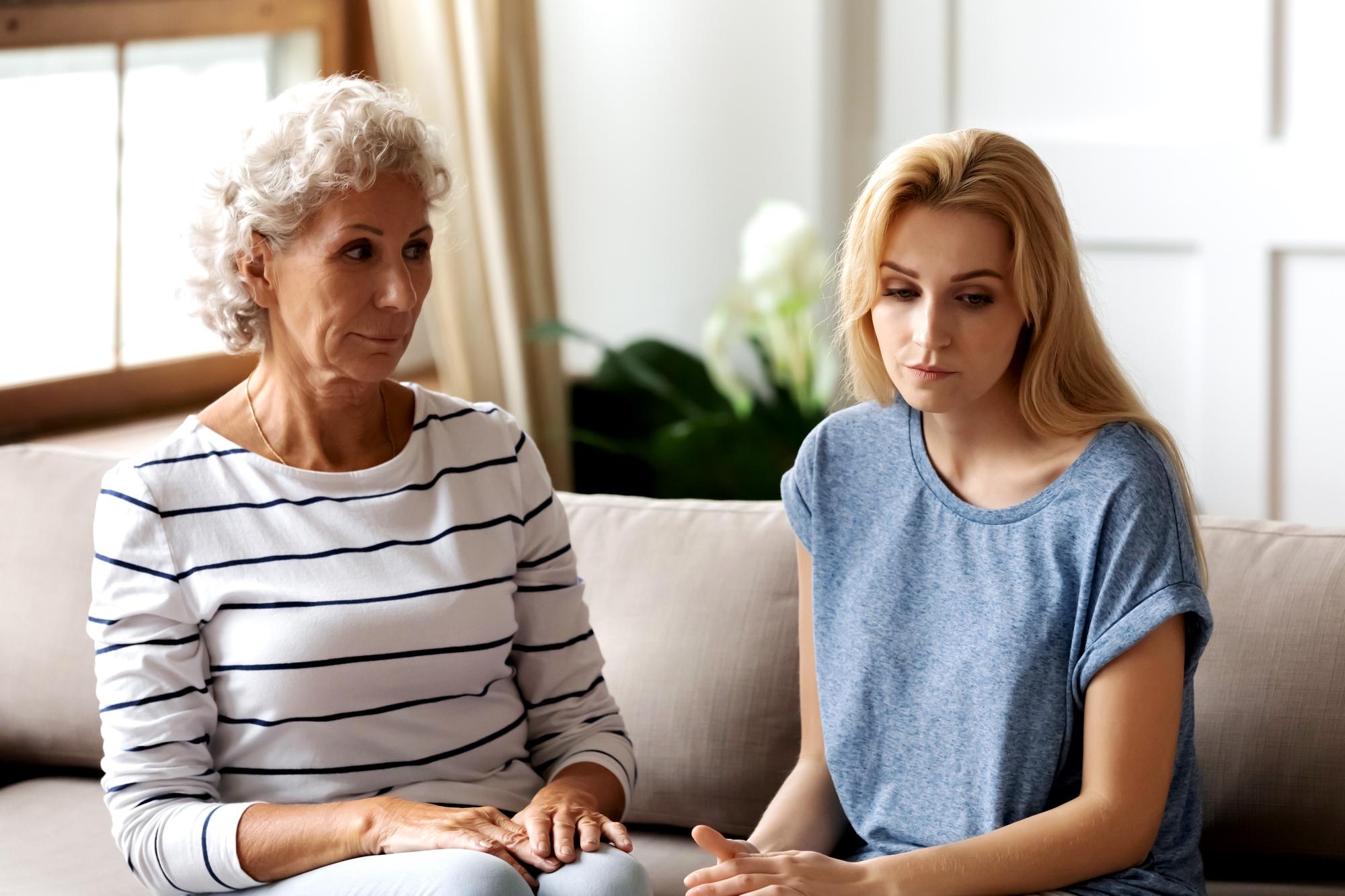 An older woman with curly gray hair and a younger woman with long blonde hair are sitting closely together on a beige sofa. Both appear to be deep in thought or concerned. The room is bright, with sunlight streaming in through a window in the background.