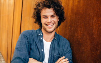 A smiling person with curly hair leans against a rust-colored wall. They are wearing a blue denim shirt over a white t-shirt and have their arms crossed. The background includes additional rust-colored panels extending to the left.