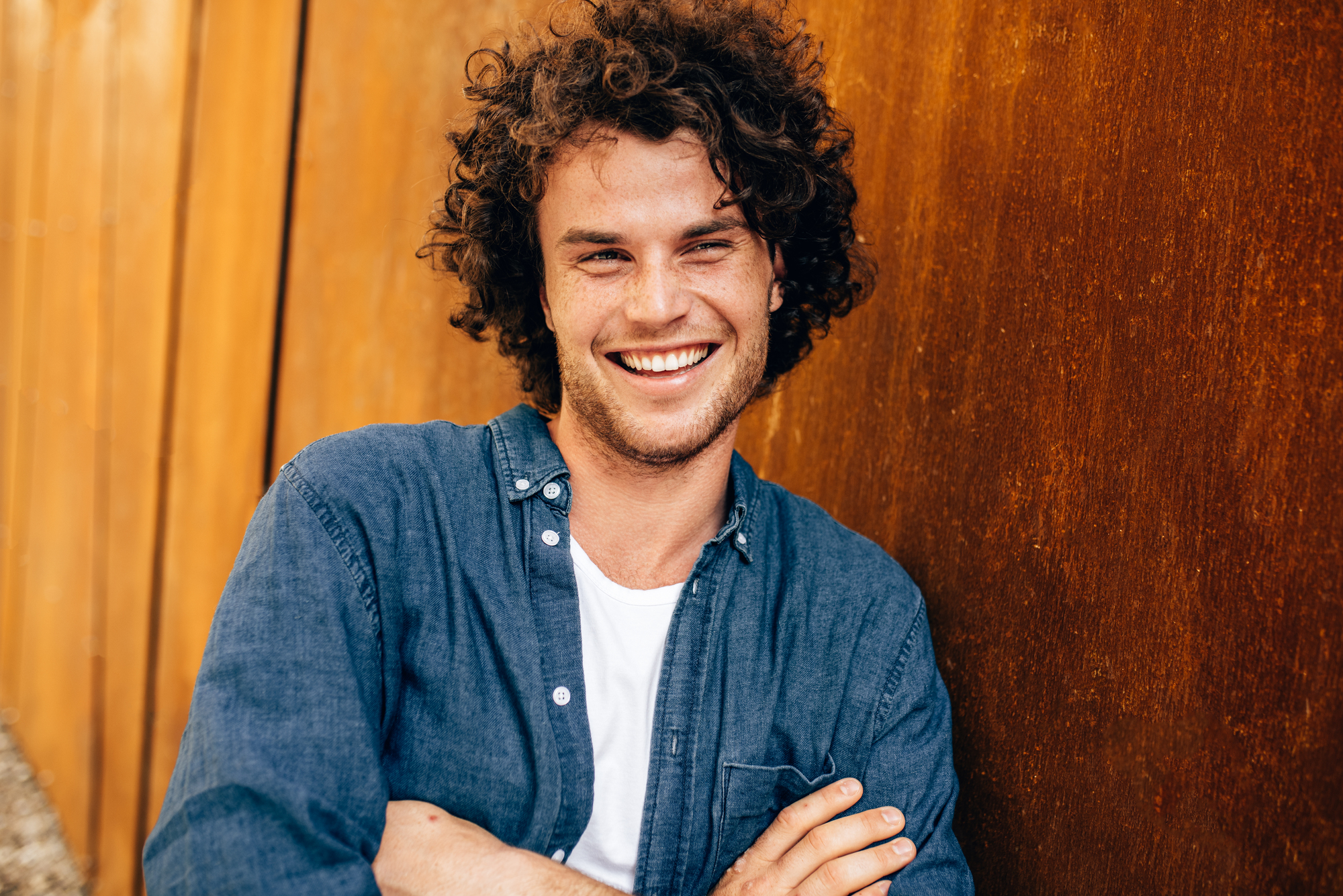 A smiling person with curly hair leans against a rust-colored wall. They are wearing a blue denim shirt over a white t-shirt and have their arms crossed. The background includes additional rust-colored panels extending to the left.