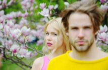 A woman with long blonde hair wearing a pink top stands among blooming magnolia flowers, gazing at the camera. A man with tousled light brown hair and a beard wearing a yellow sweater stands in the foreground, slightly out of focus.