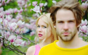 A woman with long blonde hair wearing a pink top stands among blooming magnolia flowers, gazing at the camera. A man with tousled light brown hair and a beard wearing a yellow sweater stands in the foreground, slightly out of focus.