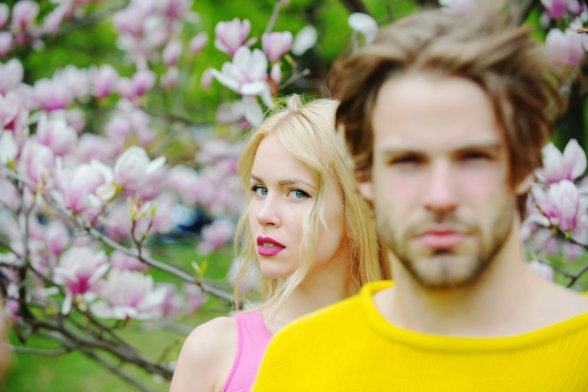 A woman with long blonde hair wearing a pink top stands among blooming magnolia flowers, gazing at the camera. A man with tousled light brown hair and a beard wearing a yellow sweater stands in the foreground, slightly out of focus.