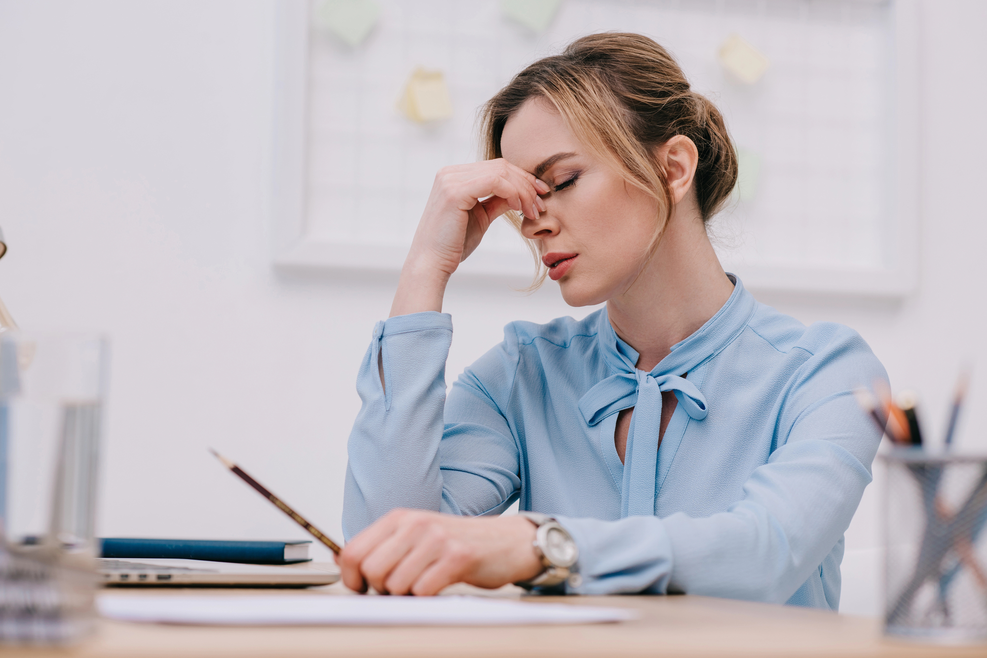 A woman wearing a light blue blouse sits at a desk with her eyes closed, pinching the bridge of her nose in frustration or fatigue. There are papers, a pencil, a laptop, and office supplies on the desk. The background shows a whiteboard with sticky notes.