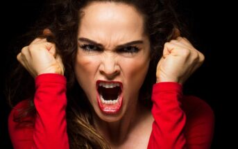 A woman with curly hair wearing a red top is screaming while clenching her fists and showing an angry facial expression against a black background.