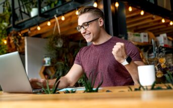 A man wearing glasses and a maroon t-shirt is sitting at a wooden table, looking at a laptop screen and raising his fist in excitement. The background shows shelves with plants and hanging lights. A white coffee mug and a notebook are also on the table.