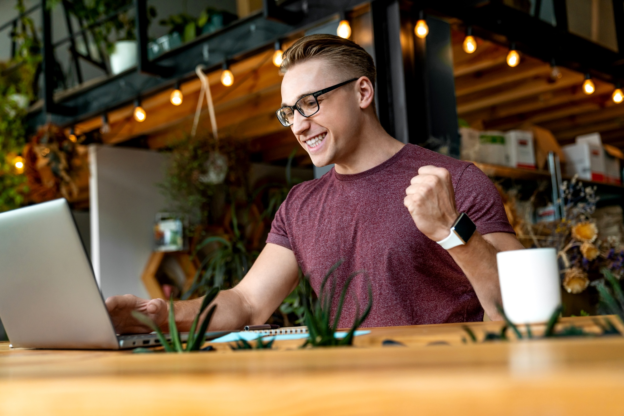 A man wearing glasses and a maroon t-shirt is sitting at a wooden table, looking at a laptop screen and raising his fist in excitement. The background shows shelves with plants and hanging lights. A white coffee mug and a notebook are also on the table.