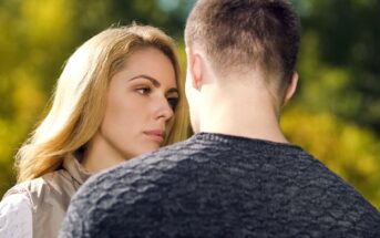 A woman with long blonde hair gazes intently at a man whose back is turned towards the camera. They stand close together in an outdoor setting with green foliage blurred in the background. The scene suggests a serious or emotional conversation.