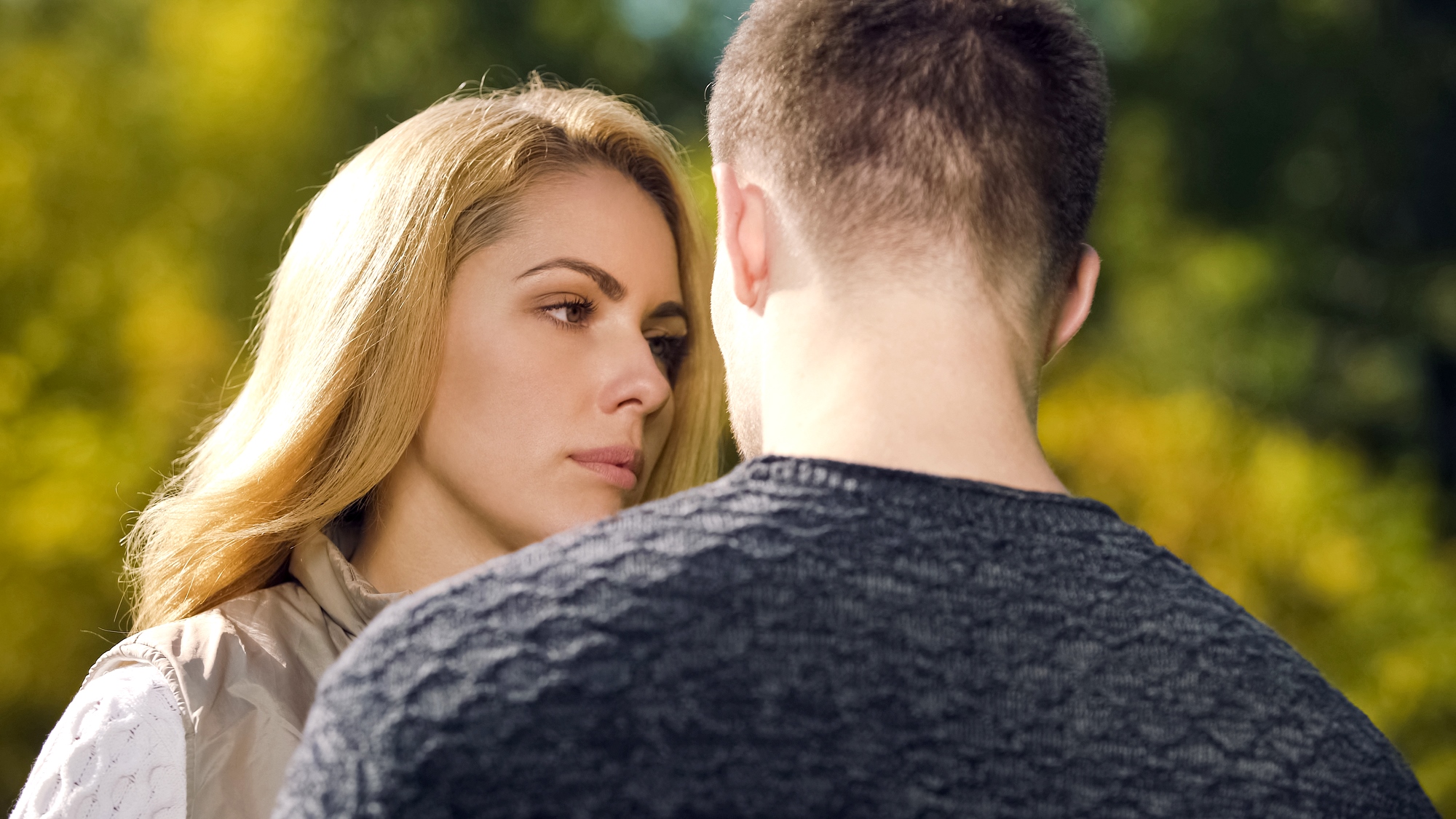 A woman with long blonde hair gazes intently at a man whose back is turned towards the camera. They stand close together in an outdoor setting with green foliage blurred in the background. The scene suggests a serious or emotional conversation.