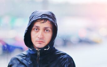 A man wearing a black hooded rain jacket stands outside in the rain. His hood is up, with wet hair peeking out. The expression on his face is neutral or slightly solemn. The blurred background suggests a rainy urban setting.