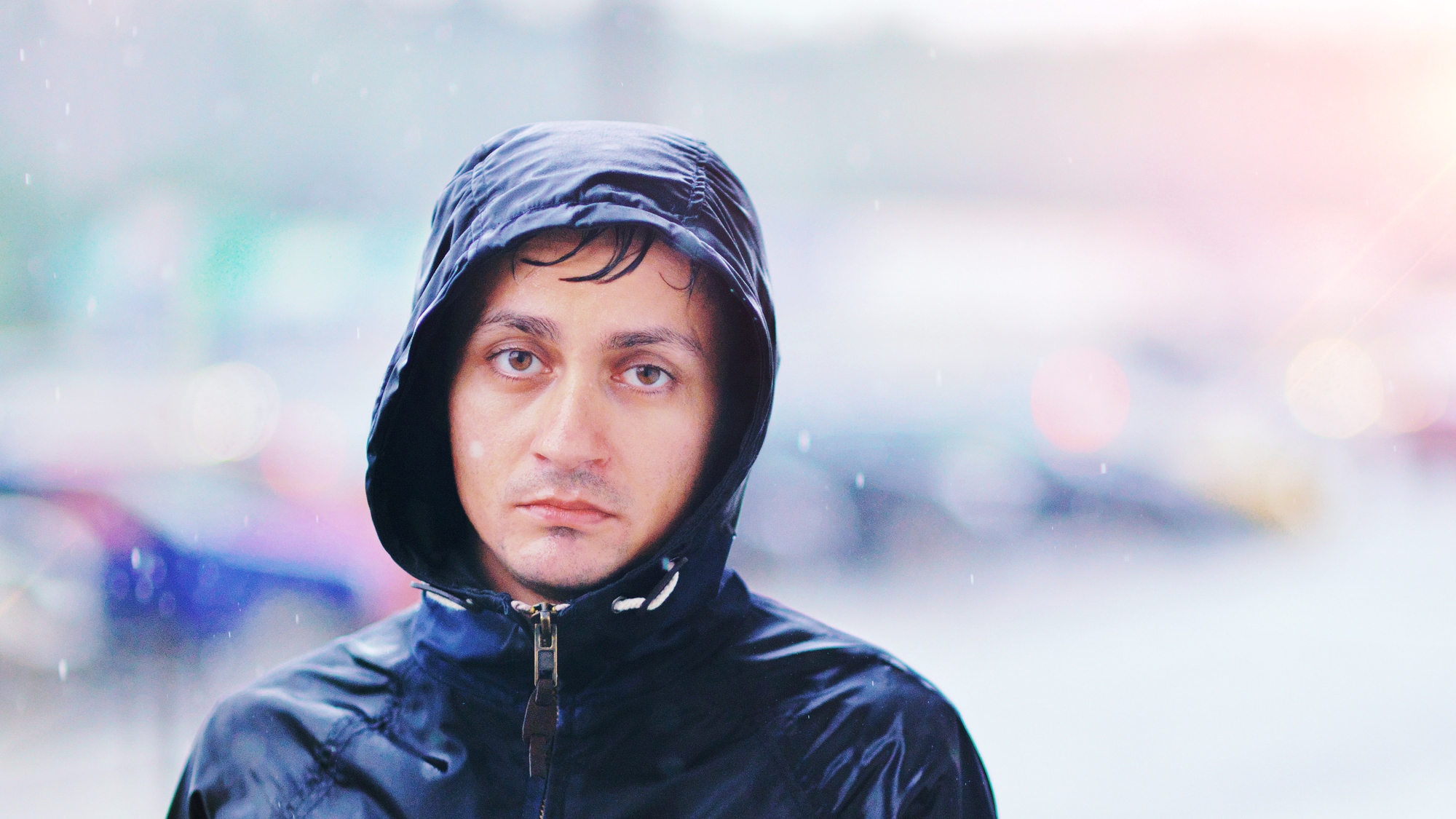 A man wearing a black hooded rain jacket stands outside in the rain. His hood is up, with wet hair peeking out. The expression on his face is neutral or slightly solemn. The blurred background suggests a rainy urban setting.