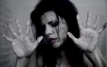 A person with dark hair, wet and disheveled, presses their hands against a foggy glass surface, their expression intense and somber. The background is blurred, adding to the moody and emotional atmosphere of the image.